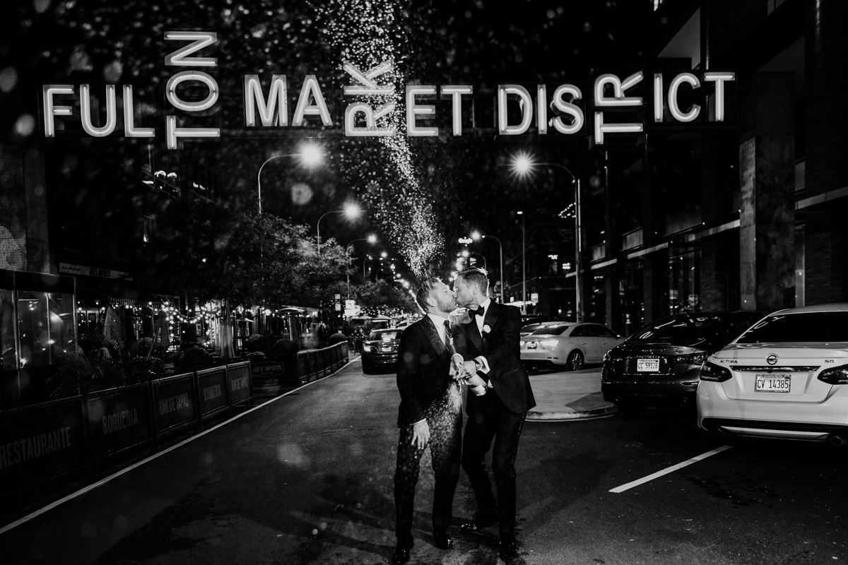Black and white photo of two grooms popping champagne under Fulton Market District sign at night