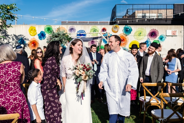 Happy bride and groom exit their summer Jewish wedding ceremony at Ignite Glass Studio in Chicago while guests cheer