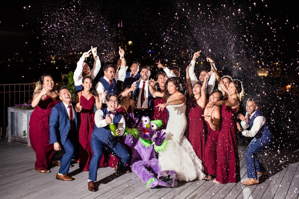 Fun wedding party photo of bride and groom popping champagne on rooftop of Lacuna Lofts with wedding party and purple creature costume