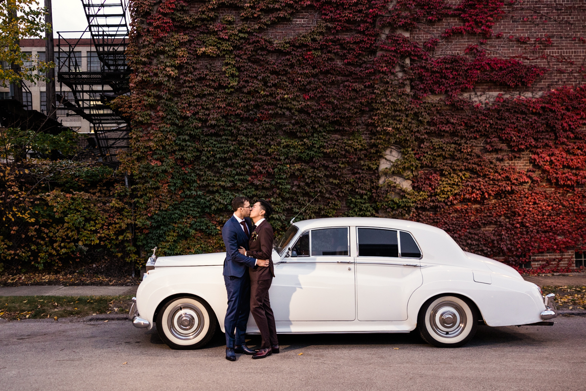 Two grooms kiss in front of classic Rolls Royce and ivy wall before fall wedding reception at Ovation Chicago