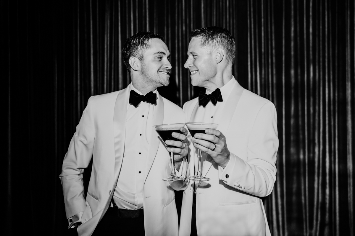 Two grooms cheers espresso martinis while wearing white tuxedo jackets at their downtown Chicago wedding