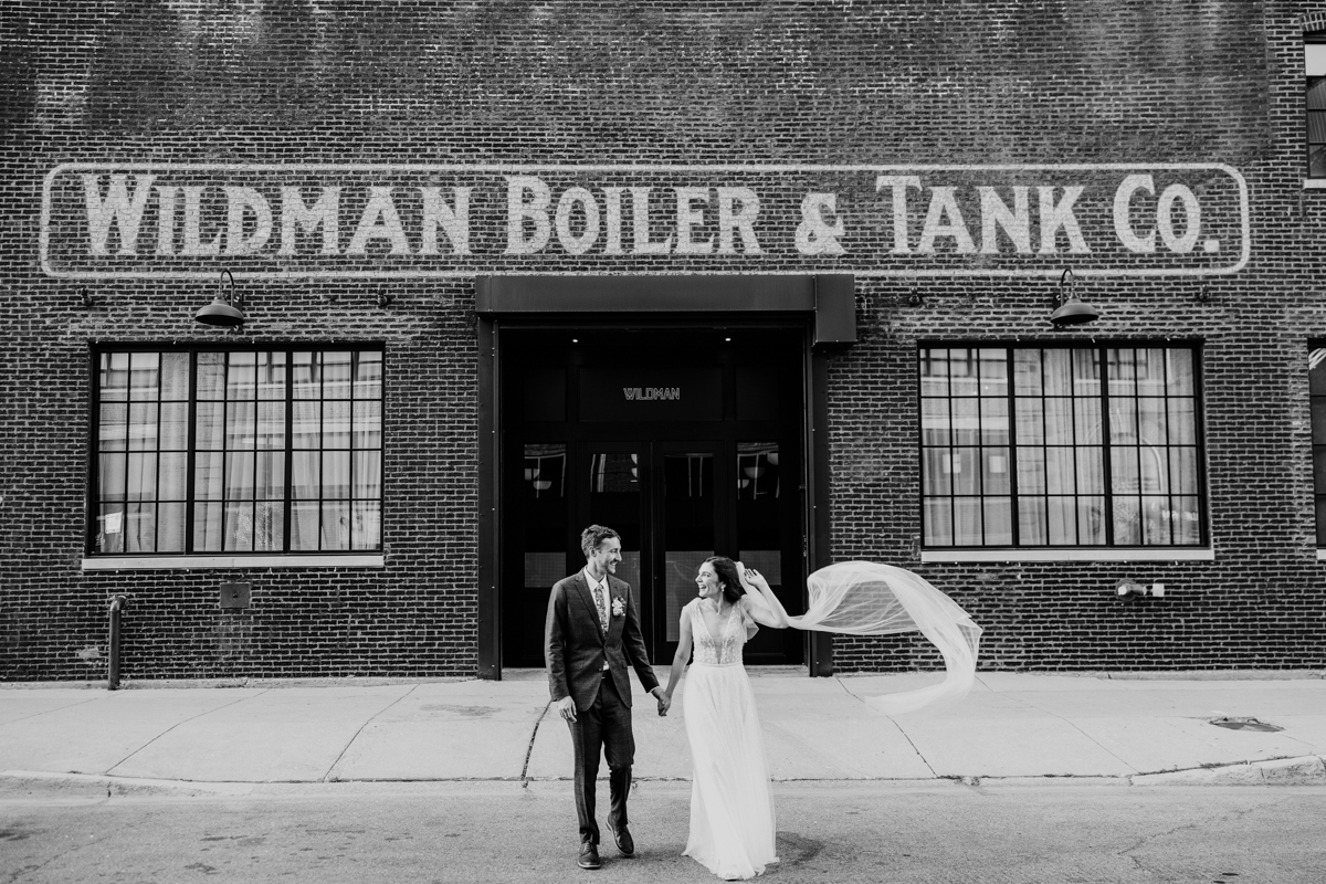 Black and white photo of bride and groom walking in front of Wildman BT wedding venue with flowing veil